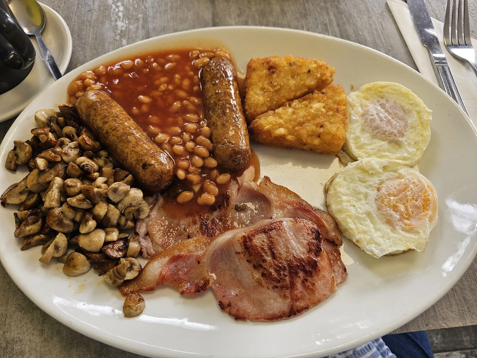 A cooked breakfast with sausages, bacon, hash browns, mushrooms, beans and two fried eggs